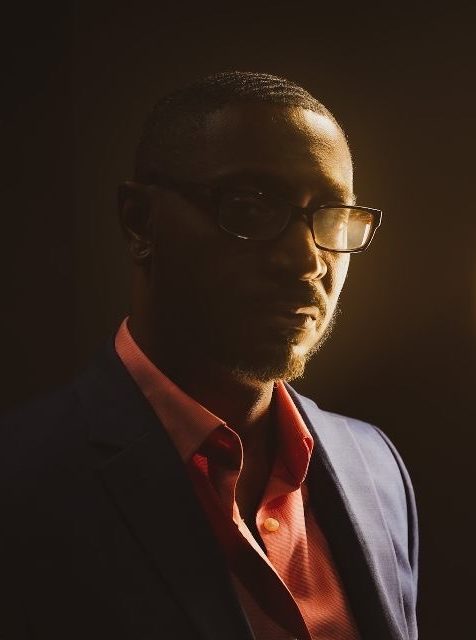 Barkeem Barr in glasses and a light suit, lit from behind in the studio